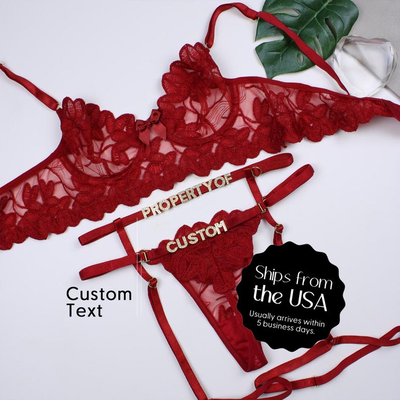 May include: A red lingerie set featuring a bra and thong, both adorned with floral lace. The set includes adjustable straps and decorative embellishments. The words "PROPERTY OF" and "CUSTOM" are displayed on the set.