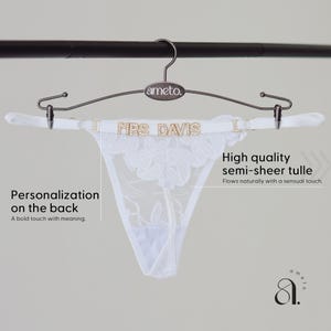 May include: White semi-sheer tulle thong underwear with floral embroidery, hanging on a metal hanger. The waistband features the text "MRS DAVIS" in gold lettering. The hanger is branded with the name "ameto."