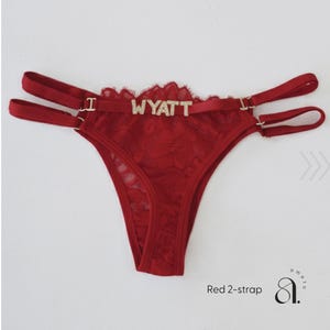 Pode incluir: Tanga de renda vermelha com al&ccedil;as duplas e o nome "WYATT" escrito em letras brilhantes. A lingerie apresenta um delicado padr&atilde;o de renda floral e uma borda ondulada. O texto "Red 2-strap" &eacute; vis&iacute;vel.