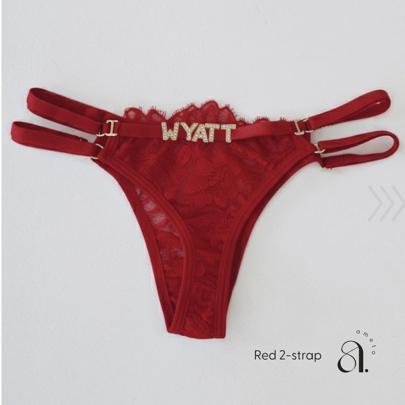 Pode incluir: Tanga de renda vermelha com al&ccedil;as duplas e o nome "WYATT" escrito em letras brilhantes. A lingerie apresenta um delicado padr&atilde;o de renda floral e uma borda ondulada. O texto "Red 2-strap" &eacute; vis&iacute;vel.
