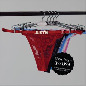 May include: A red thong with heart-shaped lace detailing, displayed on a hanger with the name "JUSTIN". Other thongs in various colors are visible behind it. The hangers are on a black rod.
