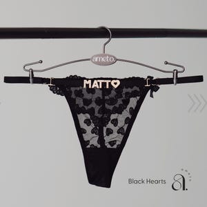 May include: Black lace thong with heart-shaped cutouts, embellished with the word "MATTO" in gold lettering. The thong is suspended from a hanger with the brand name "ameto." The text "Black Hearts" is visible.