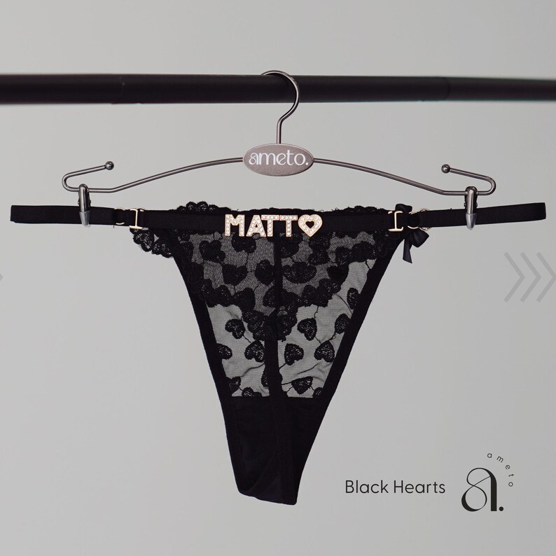 May include: Black lace thong with heart-shaped cutouts, embellished with the word "MATTO" in gold lettering. The thong is suspended from a hanger with the brand name "ameto." The text "Black Hearts" is visible.
