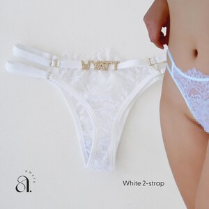 Pode incluir: Tanga branco de renda com al&ccedil;as duplas e detalhes dourados. A palavra "WYATT" est&aacute; escrita com strass na frente. A pe&ccedil;a &eacute; exibida em um fundo branco.