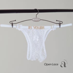 May include: White open lace thong underwear with a bow detail, displayed on a hanger with the text "IN YOURS". The hanger is silver and has the brand name "ameto" on it. The words "Open Lace" are written in the bottom left corner.