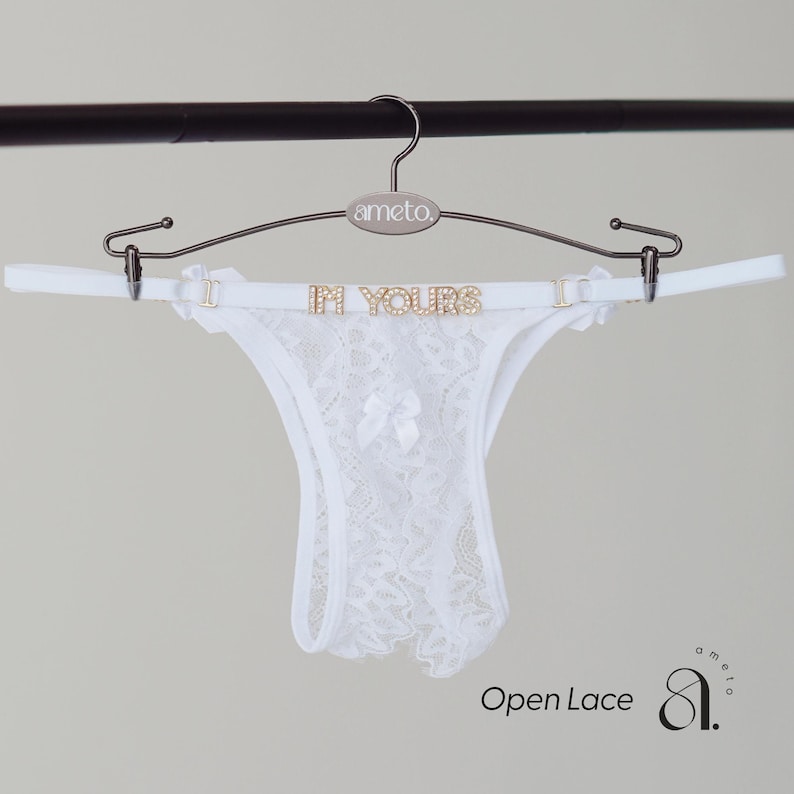 May include: White open lace thong underwear with a bow detail, displayed on a hanger with the text "IN YOURS". The hanger is silver and has the brand name "ameto" on it. The words "Open Lace" are written in the bottom left corner.