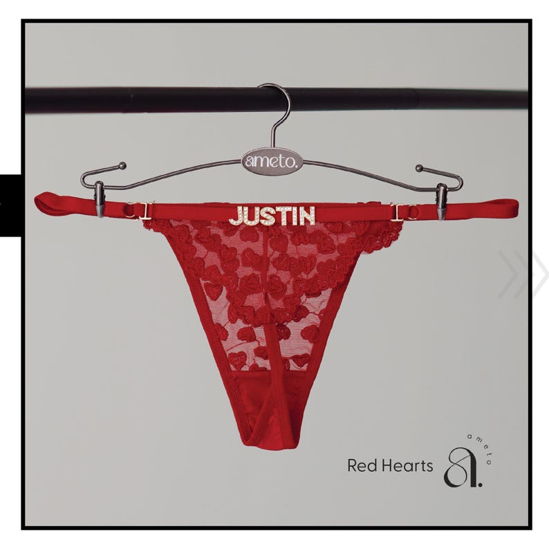May include: Red thong underwear with a heart-shaped lace design, hanging on a silver hanger. The word "JUSTIN" is spelled out in silver letters on the waistband. The text "Red Hearts" is visible.