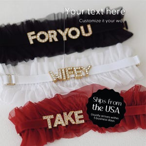 May include: Three bridal garters in black, white, and red. Each garter features a word in gold letters: "FOR YOU", "WIFEY", and "TAKE". The garters have a ruffled tulle design.