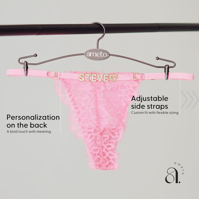 Pode incluir: Calcinha tanga rosa de renda com al&ccedil;as laterais ajust&aacute;veis, pendurada em um cabide. A cintura apresenta o nome "STEVE" em letras brilhantes. O texto diz "Personaliza&ccedil;&atilde;o nas costas".