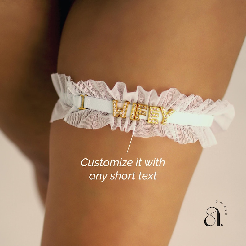 May include: A white bridal garter with the word "WIFEY" spelled out in gold letters with rhinestones. The garter has a ruffled lace trim and a light blue band. The text "Customize it with any short text" is also visible.