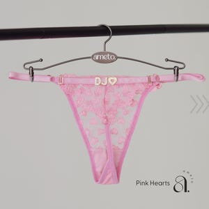 May include: Pink thong underwear with a heart-shaped lace design. The garment is suspended from a hanger with the brand name "ameto." The waistband features the letters "DJO" and a heart-shaped embellishment. The words "Pink Hearts" are visible.