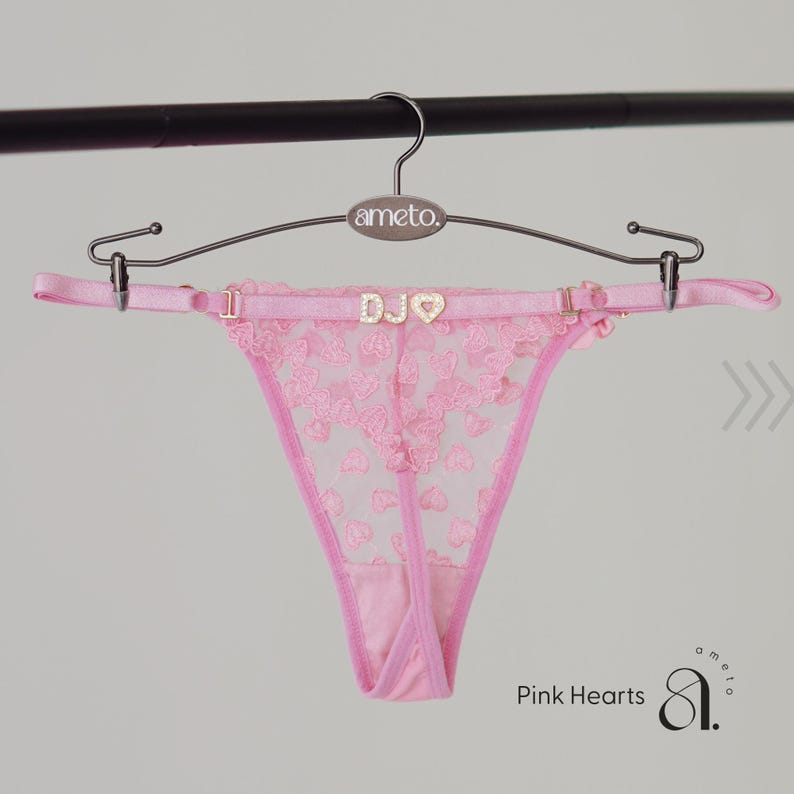 May include: Pink thong underwear with a heart-shaped lace design. The garment is suspended from a hanger with the brand name "ameto." The waistband features the letters "DJO" and a heart-shaped embellishment. The words "Pink Hearts" are visible.