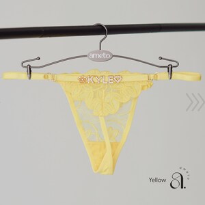 May include: A yellow thong with a floral lace design, hanging from a metal hanger. The thong features the name "KYLE" in sparkling letters and a crown. The hanger has the brand name "ameto."