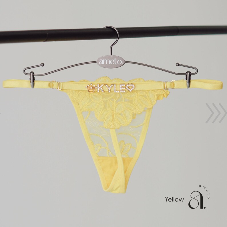 May include: A yellow thong with a floral lace design, hanging from a metal hanger. The thong features the name "KYLE" in sparkling letters and a crown. The hanger has the brand name "ameto."