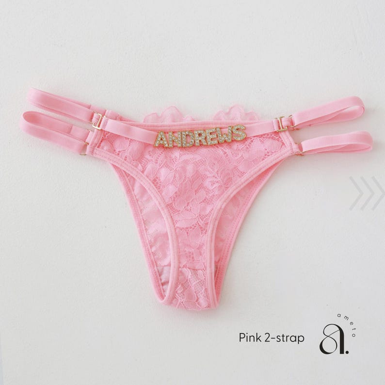Pode incluir: Tanga de renda rosa com duas al&ccedil;as e ferragens douradas. A palavra "ANDREWS" est&aacute; escrita em letras douradas na frente. O texto "Pink 2-strap" est&aacute; no canto inferior esquerdo.