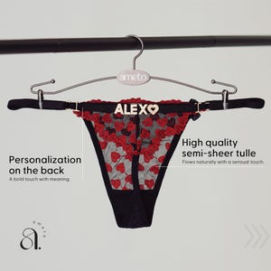 以下が含まれることがあります： 赤いハートの刺繍と金色の文字で「ALEXO」という名前が入った黒いTバック。 ランジェリーは半透明のチュールで作られており、シルバーのハンガーに吊るされています。