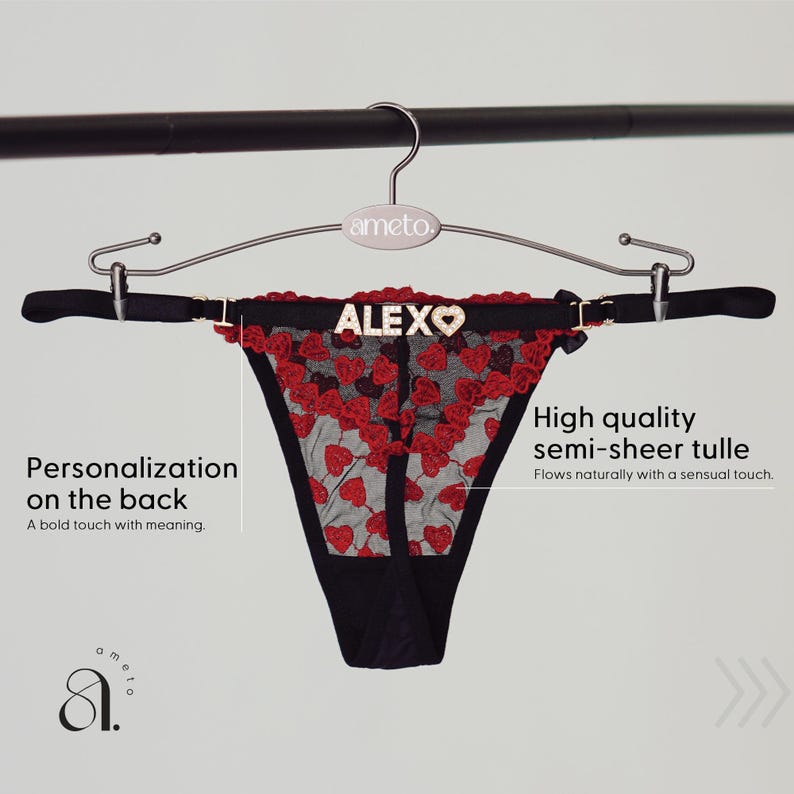 以下が含まれることがあります： 赤いハートの刺繍と金色の文字で「ALEXO」という名前が入った黒いTバック。 ランジェリーは半透明のチュールで作られており、シルバーのハンガーに吊るされています。
