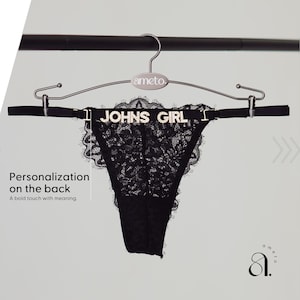 Pode incluir: Calcinha fio dental de renda preta com o texto "JOHNS GIRL" em letras prateadas. A roupa &iacute;ntima &eacute; exibida em um cabide prateado com o nome da marca "ameto". A imagem tamb&eacute;m inclui o texto "Personaliza&ccedil;&atilde;o nas costas. Um toque ousado com significado."