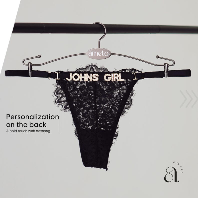 Pode incluir: Calcinha fio dental de renda preta com o texto "JOHNS GIRL" em letras prateadas. A roupa &iacute;ntima &eacute; exibida em um cabide prateado com o nome da marca "ameto". A imagem tamb&eacute;m inclui o texto "Personaliza&ccedil;&atilde;o nas costas. Um toque ousado com significado."