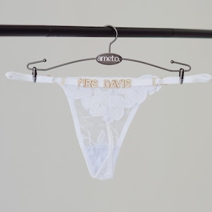 May include: A white lace thong with floral detailing is displayed on a hanger. The thong features the text "MRS DAVIS" in gold lettering. The hanger is a dark metal color and has the brand name "ameto" printed on it.