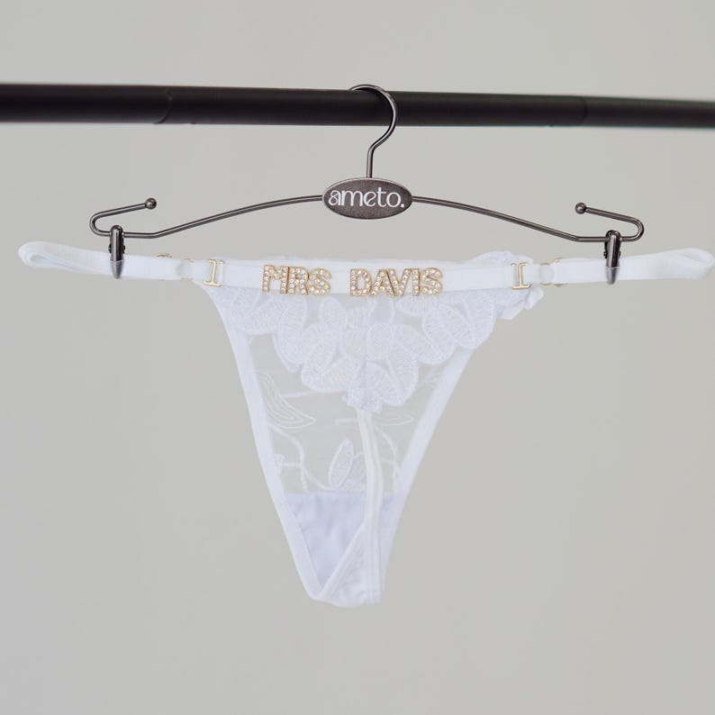 May include: A white lace thong with floral detailing is displayed on a hanger. The thong features the text "MRS DAVIS" in gold lettering. The hanger is a dark metal color and has the brand name "ameto" printed on it.