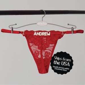 May include: Red lace thong underwear with the name "ANDREW" spelled out in silver letters. The underwear is hanging on a silver hanger with the brand name "ameto" on it.