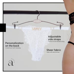 Pode incluir: Calcinha tanga de renda branca com a palavra "Wifey" em letras douradas na cintura, pendurada em um cabide prateado. A calcinha possui al&ccedil;as laterais ajust&aacute;veis e tecido transparente.