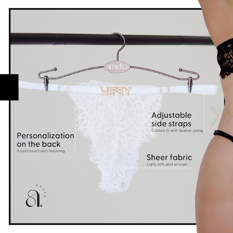 Pode incluir: Calcinha tanga de renda branca com a palavra "Wifey" em letras douradas na cintura, pendurada em um cabide prateado. A calcinha possui al&ccedil;as laterais ajust&aacute;veis e tecido transparente.
