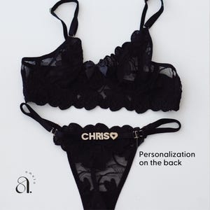 May include: Black lace lingerie set featuring a bralette and thong. The bralette has adjustable straps and a bow detail. The thong has the name "CHRIS" spelled out in rhinestones with a heart symbol. The set is displayed on a white background.