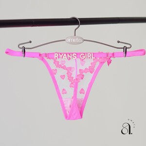 以下が含まれることがあります： 鮮やかなピンク色のTバック。ハート模様と「RYANS GIRL」の文字がキラキラと輝いています。ランジェリーは「ameto」のブランド名が入ったハンガーにかけられています。