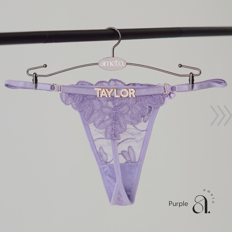 May include: A lavender thong with floral lace detailing, displayed on a hanger. The word "TAYLOR" is spelled out in gold letters across the waistband. The hanger is silver and has the brand name "ameto" printed on it.
