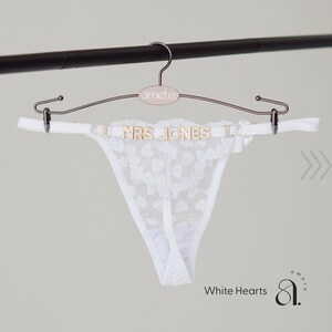 May include: White lace thong with heart patterns, displayed on a rose gold hanger. The hanger features the text "I MRS JONES I" in gold lettering. The hanger is suspended from a black bar. The text "White Hearts" is visible.