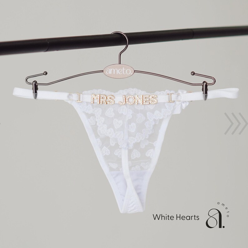 May include: White lace thong with heart patterns, displayed on a rose gold hanger. The hanger features the text "I MRS JONES I" in gold lettering. The hanger is suspended from a black bar. The text "White Hearts" is visible.