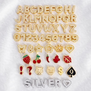 May include: A collection of gold-colored alphabet letters, numbers, and various charms, all encrusted with small, sparkling stones. The charms include a strawberry, cherries, a chili pepper, and a cat face.