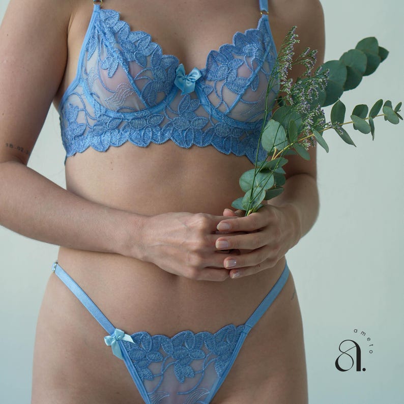 May include: Light blue lingerie set featuring a floral lace design. The set includes a bra with scalloped edges and a matching thong, both adorned with small bows. The bra has adjustable straps. The lingerie is made of a sheer, delicate fabric.