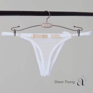 May include: White sheer thong underwear with the text "JESSE'S GIRL" in gold rhinestones. The thong is hanging on a silver hanger. The words "Sheer Thong" are printed in black at the bottom of the image.
