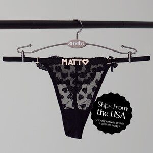 May include: Black thong underwear with a heart-shaped lace pattern. The waistband features the name "MATTO" in gold letters and a small bow. The underwear is displayed on a hanger.