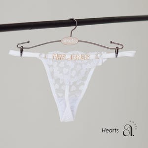 May include: White lace thong underwear with a heart pattern, hanging on a rose gold hanger. The hanger has the word "ameto" on it. The thong has the text "MRS JONES" in gold letters.