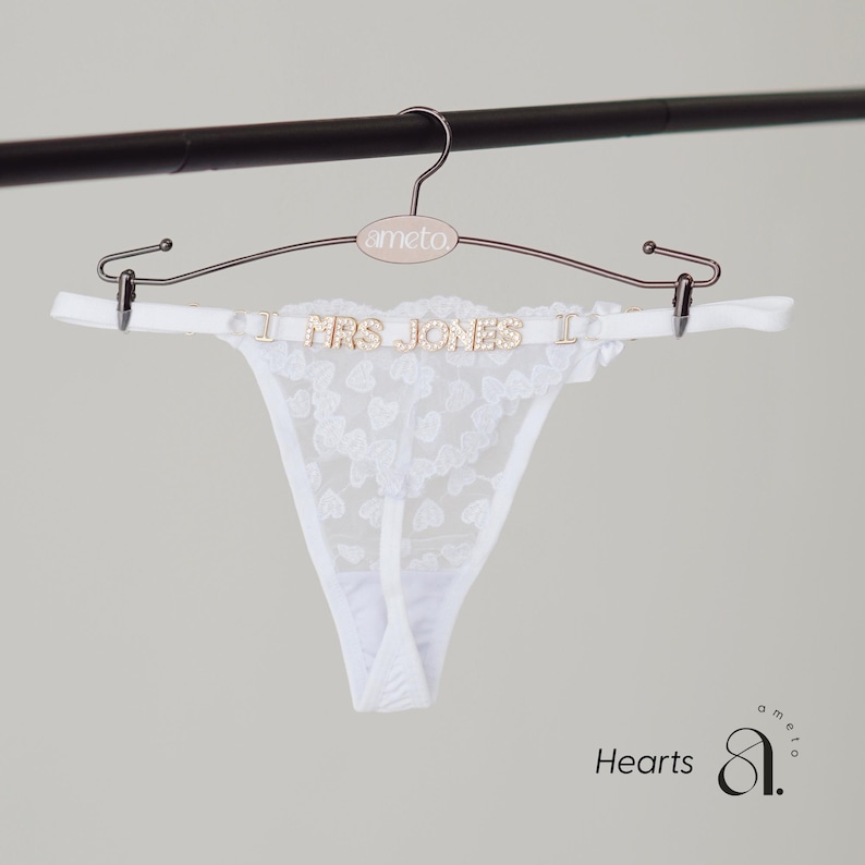 May include: White lace thong underwear with a heart pattern, hanging on a rose gold hanger. The hanger has the word "ameto" on it. The thong has the text "MRS JONES" in gold letters.
