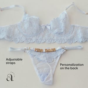 May include: Light blue lingerie set featuring a bra and thong. The bra has a floral lace design and a small bow. The thong has the text "MRS DAVIS" in gold letters. Adjustable straps are visible. The set is on a white background.