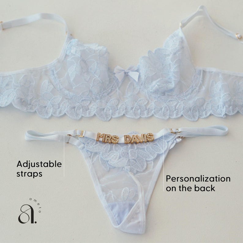 May include: Light blue lingerie set featuring a bra and thong. The bra has a floral lace design and a small bow. The thong has the text "MRS DAVIS" in gold letters. Adjustable straps are visible. The set is on a white background.