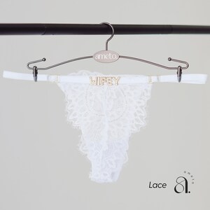 May include: White lace thong underwear with the word "Wifey" spelled out in gold-colored letters. The underwear is hanging on a silver hanger with the brand name "ameto". The word "Lace" is written in the bottom left corner.