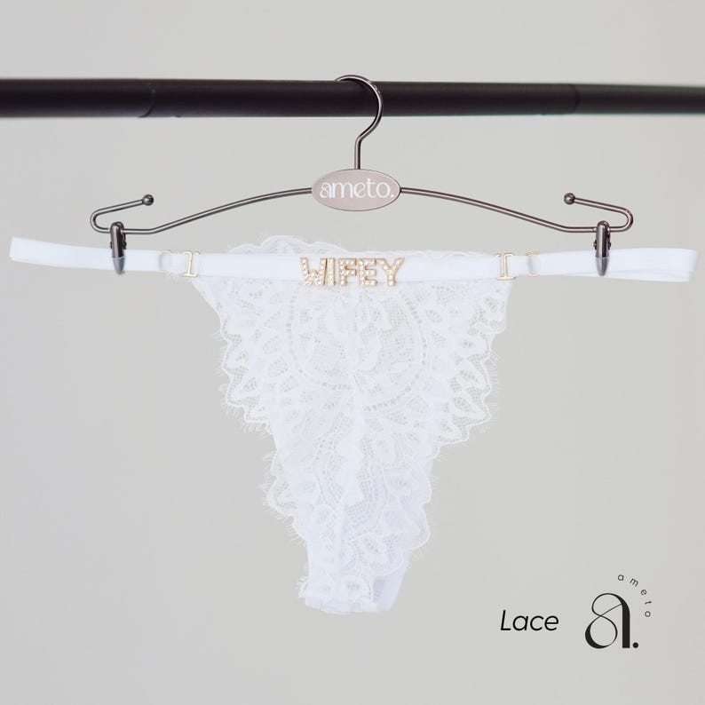 May include: White lace thong underwear with the word "Wifey" spelled out in gold-colored letters. The underwear is hanging on a silver hanger with the brand name "ameto". The word "Lace" is written in the bottom left corner.