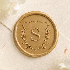 May include: A circular gold wax seal featuring a central letter S embossed within a shield-like outline, surrounded by a delicate laurel leaf wreath design. The seal is pressed onto a textured cream-colored envelope surface.
