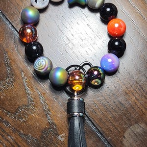 May include: A black leather tassel keychain with a silver ring and a charm made of iridescent beads, including a star, a swirl, and a black bead with a white bat design.