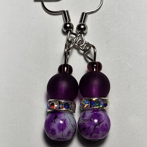 May include: A pair of dangle earrings with silver-colored ear wires. Each earring features a dark purple bead, a clear bead with iridescent rhinestones, and a white and purple patterned bead. The earrings are designed for pierced ears.