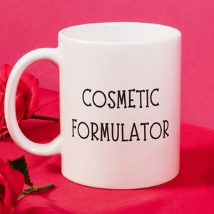 May include: White ceramic mug with the text "COSMETIC FORMULATOR" in black lettering. The mug is sitting on a pink surface with red roses scattered around it.