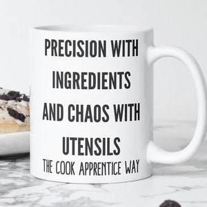 May include: White ceramic mug with black text that reads "Precision with ingredients and chaos with utensils The cook apprentice way".