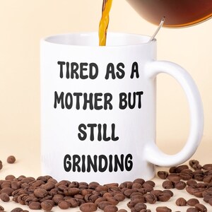 May include: A white ceramic mug with the text "TIRED AS A MOTHER BUT STILL GRINDING" is being filled with coffee. Coffee beans are scattered around the base of the mug. A clear glass carafe pours the coffee.