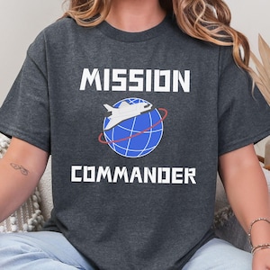 May include: Dark heather grey t-shirt with the words "MISSION COMMANDER" in white block letters. A white airplane graphic is above a blue globe with red lines. The text "DARK HEATHER GREY" is at the bottom.
